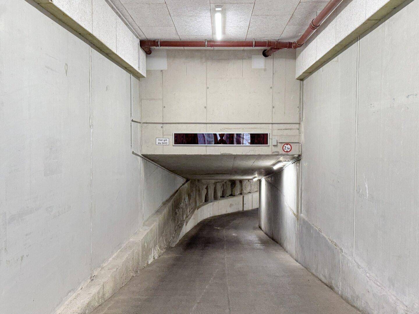 Einfahrt zur Tiefgarage mit Betonwänden, einer Höhenbegrenzung und sichtbaren Rohrleitungen.