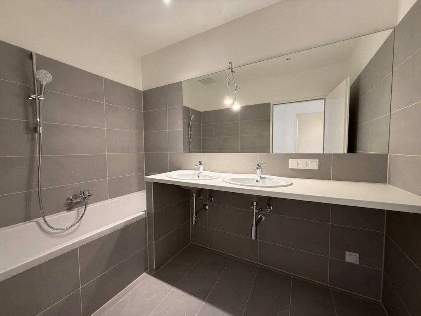 Modernes Badezimmer mit Doppelwaschbecken, Badewanne mit Duschfunktion und großen grauen Fliesen.