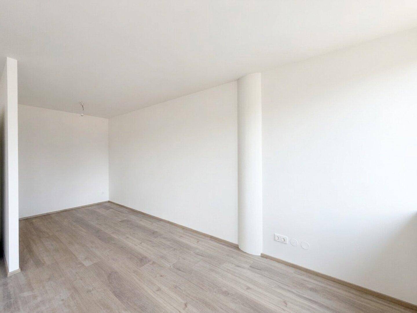 Eine Ecke des hellen Wohnraums mit weiß gestrichenen Wänden und modernem Laminatboden, bereit zur individuellen Gestaltung.