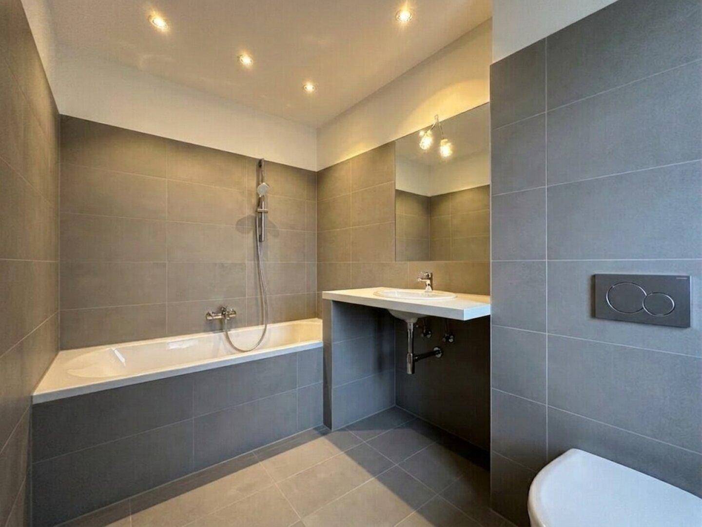 Modernes Badezimmer mit grauen Großformatfliesen, Badewanne mit Duschfunktion und wandhängendem WC.