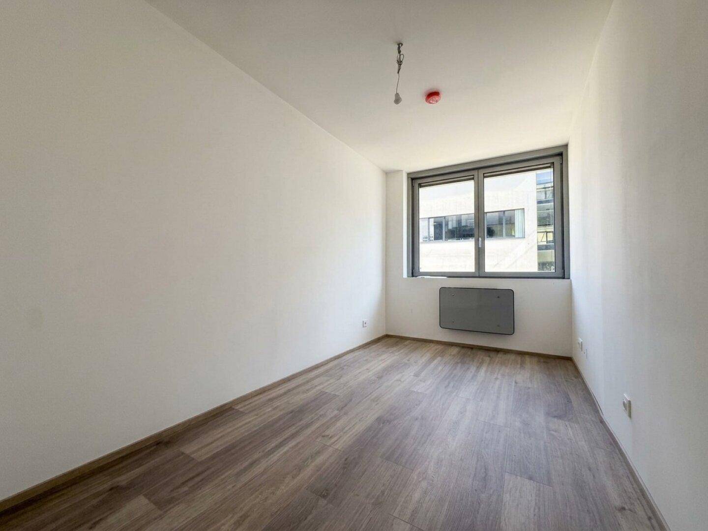Unmöbliertes Zimmer mit Laminatboden und großem Fenster, bereit für den Erstbezug in einem Neubau.