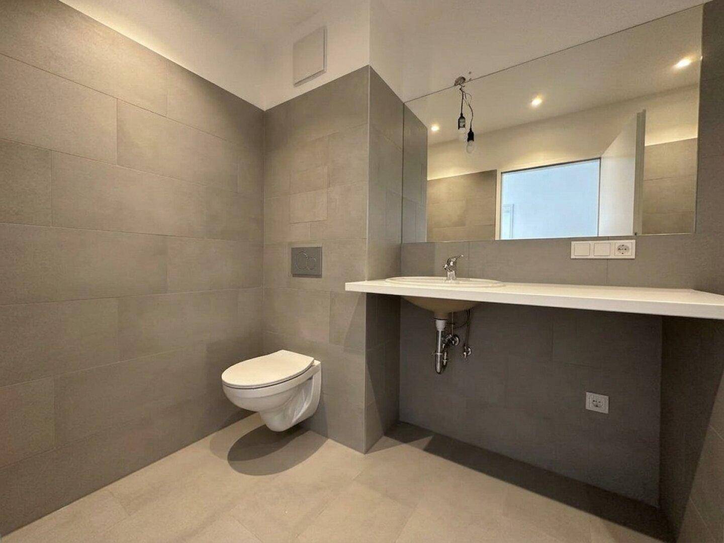 Modernes Badezimmer mit grauen Fliesen, wandhängendem WC, Einzelwaschbecken und großem Spiegel.