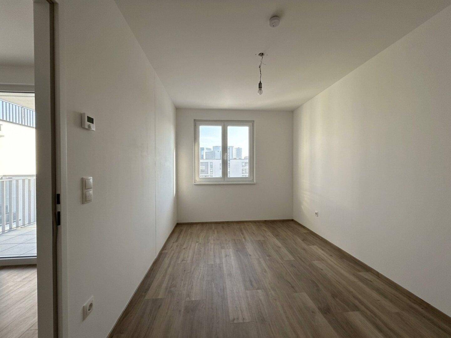 Helles Schlafzimmer mit Holzboden und Fenster mit Blick auf die Stadt, unmöbliert.