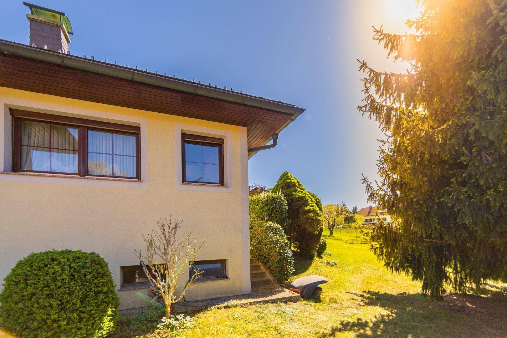 Seitenansicht des Hauses mit heller Fassade und Fenstern, umgeben von grüner Natur.