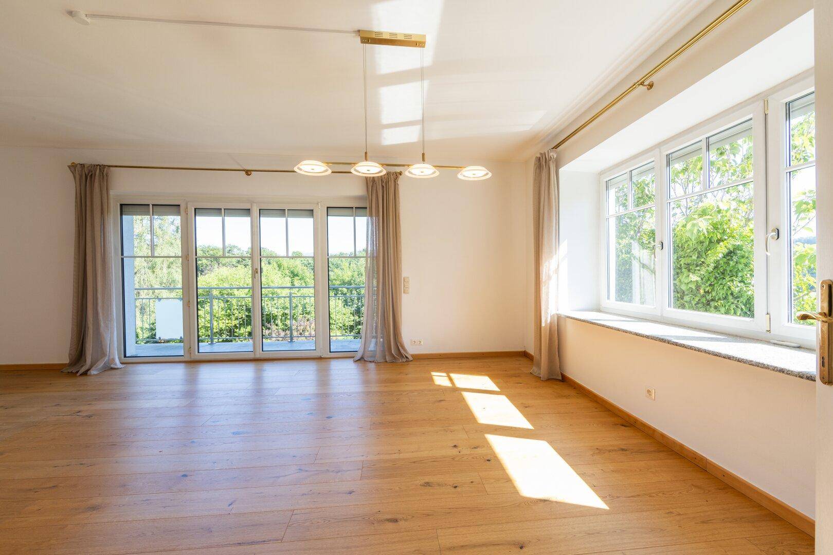 Helles, leeres Wohnzimmer mit Holzboden, großen Fenstern und Zugang zum Balkon.