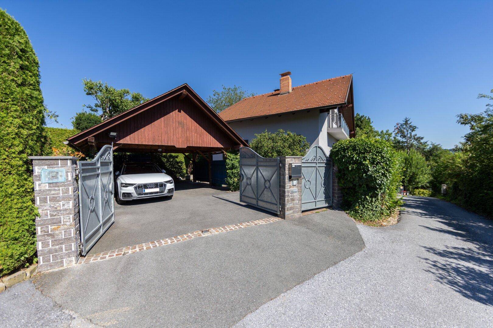 Einfahrt zum Haus mit Carport, elektrischem Tor und gepflegter Hecke unter blauem Himmel.