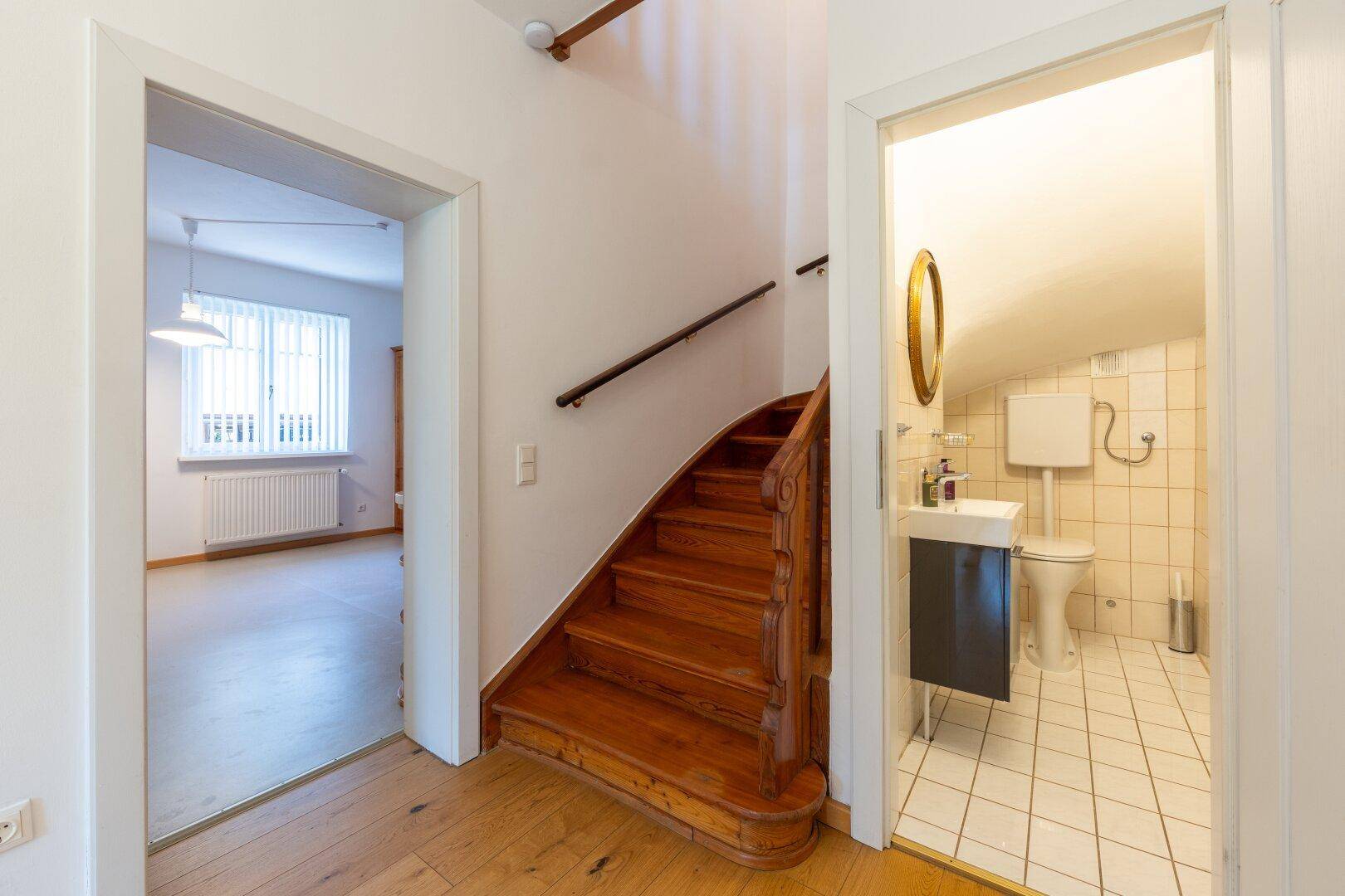 Flur mit Holztreppe und Blick in ein kleines Badezimmer mit WC und Waschbecken.