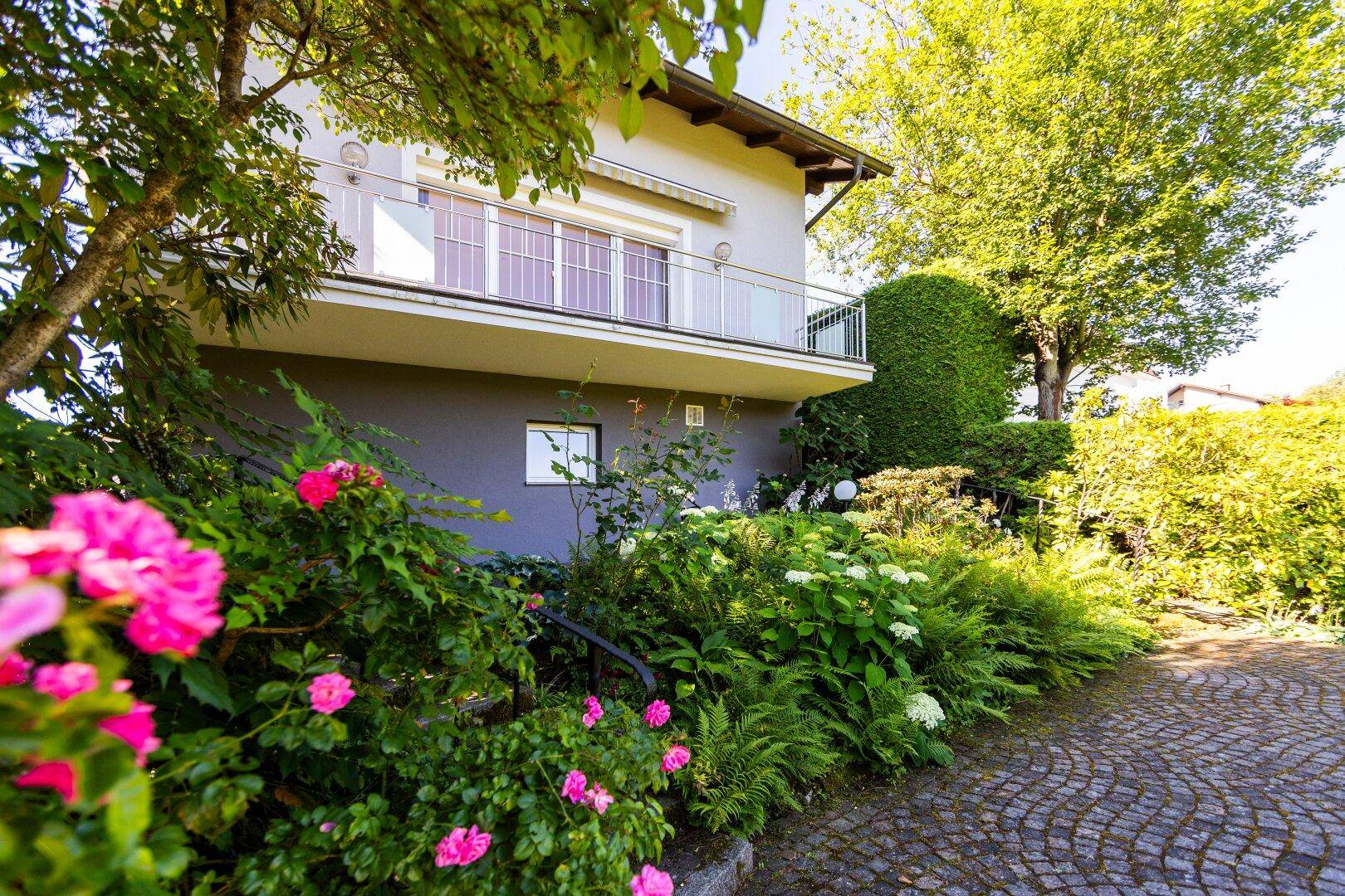 Seitenansicht des Hauses mit Balkon, üppiger Bepflanzung und gepflastertem Weg.