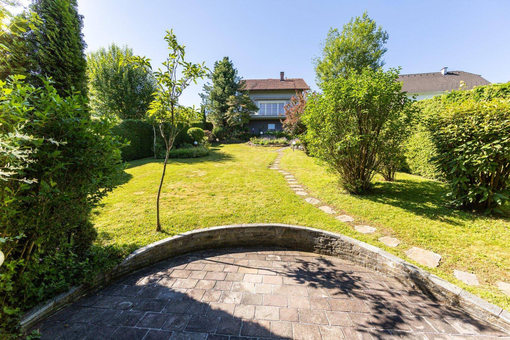 Großer, sonniger Garten mit Rasenfläche, gepflasterter Terrasse und umgebenden Bäumen.