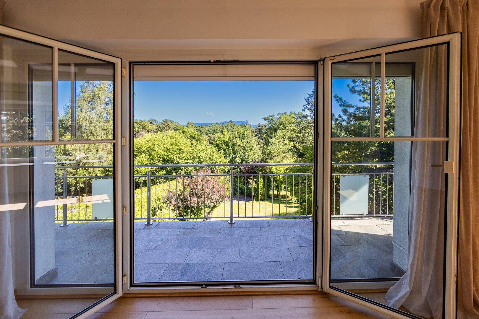 Blick vom Innenraum auf den gefliesten Balkon mit Metallgeländer und weitreichender grüner Aussicht.