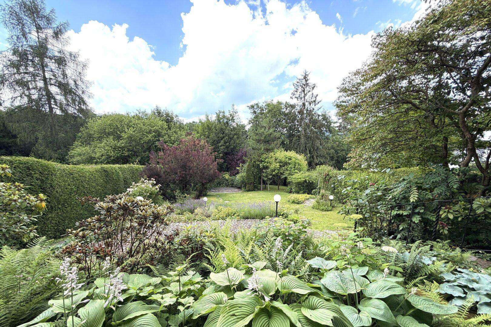 Weitläufiger Garten mit vielfältiger Bepflanzung, Rasenflächen und hohen Bäumen unter blauem Himmel.