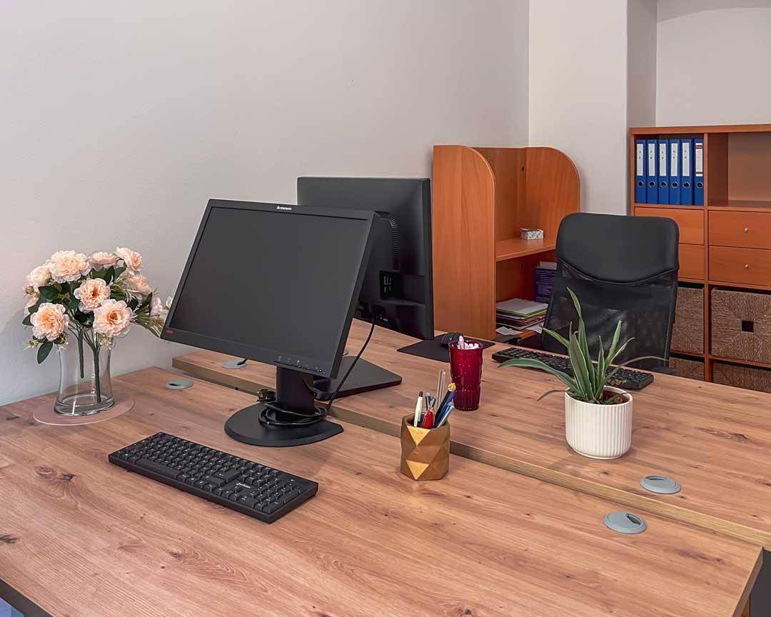 Arbeitsplätze mit Holzschreibtischen, Computern und einem Regal mit Aktenordnern in einem Büro.