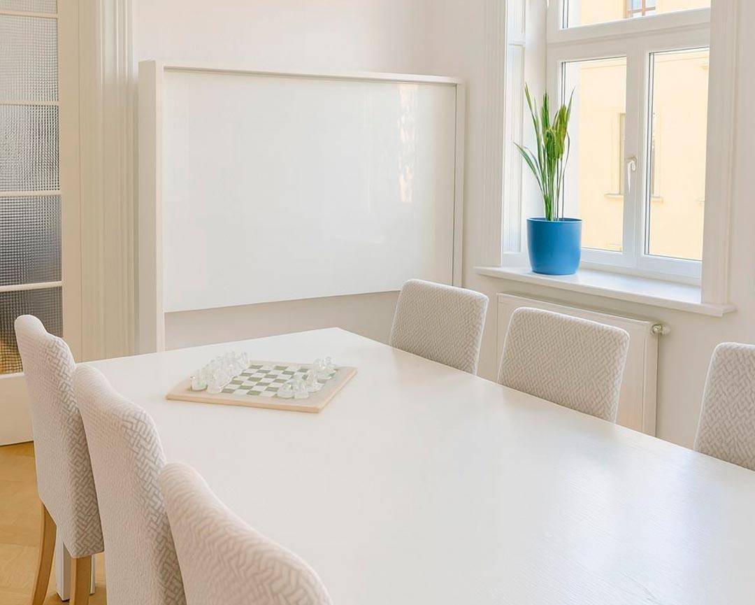 Heller Besprechungsraum mit großem weißen Tisch, bequemen Stühlen und einem Fenster mit Topfpflanze.