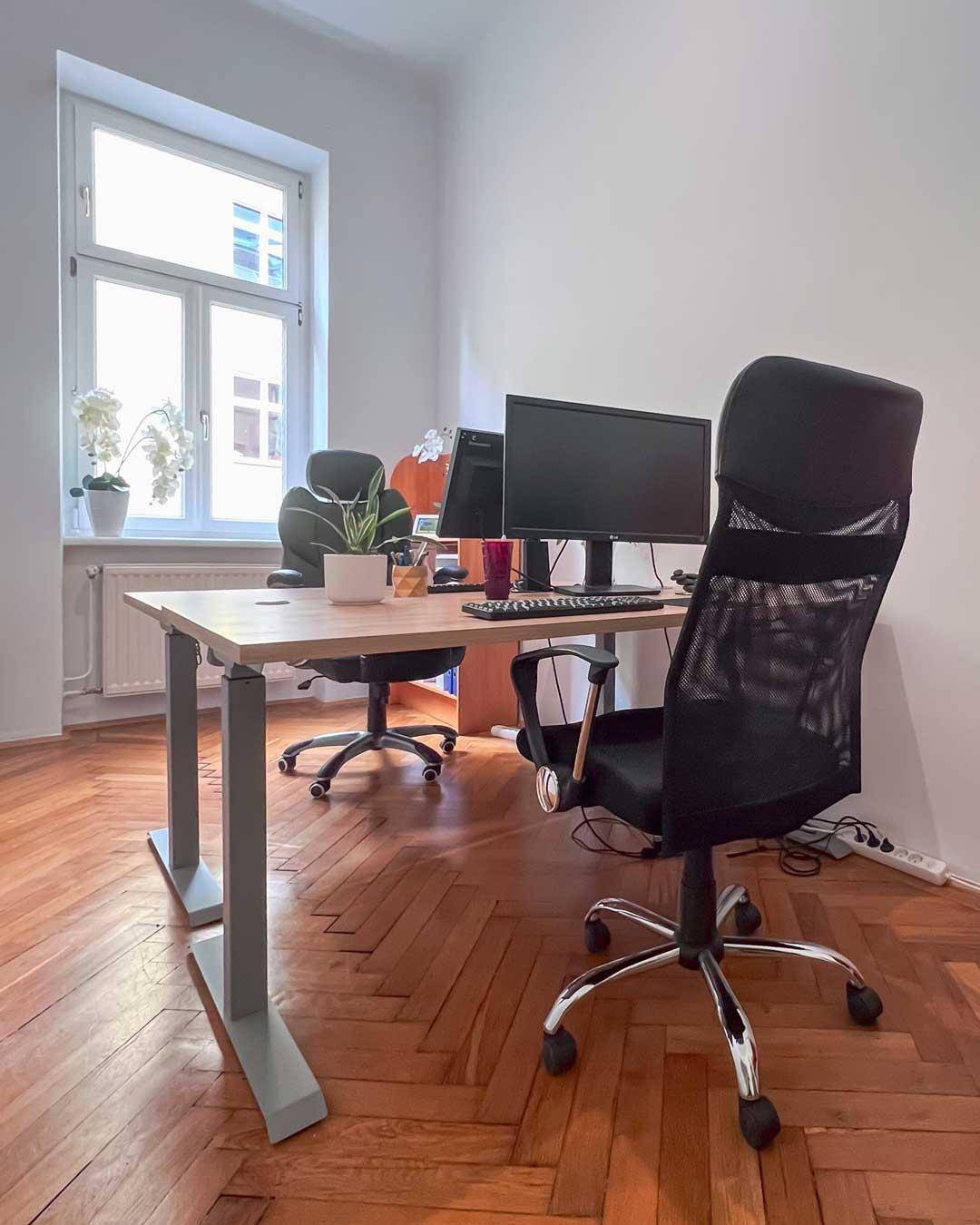 Helles Büro mit zwei modernen Arbeitsplätzen, Computern und einem großen Fenster, ausgestattet mit Parkettboden.