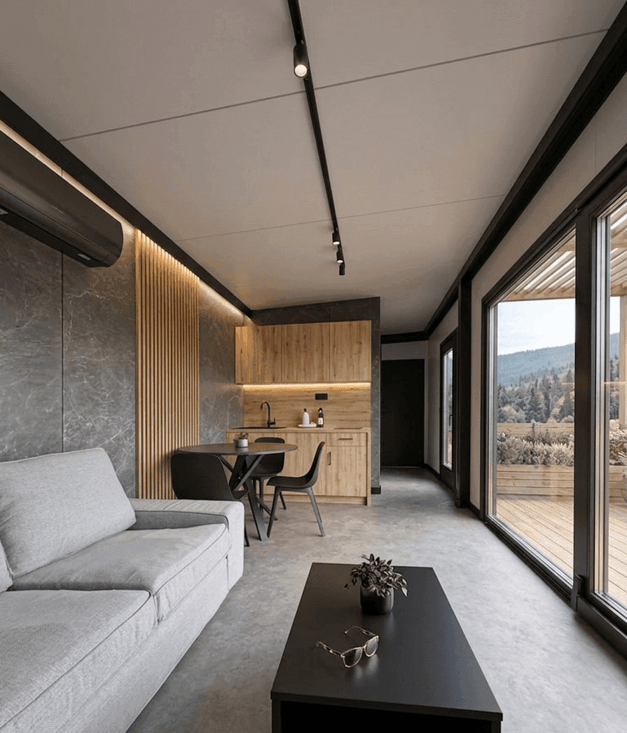 Offener Wohnbereich mit Sofa, kleiner Küchenzeile und großen Fenstern mit Zugang zur Terrasse.