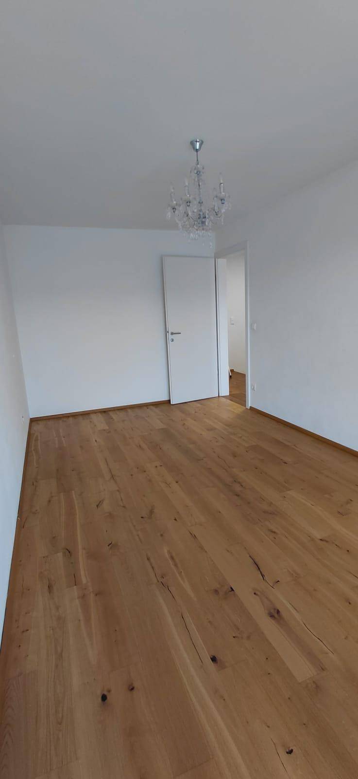 Leeres Zimmer mit hellem Parkettboden, weißen Wänden und einem eleganten Kronleuchter.