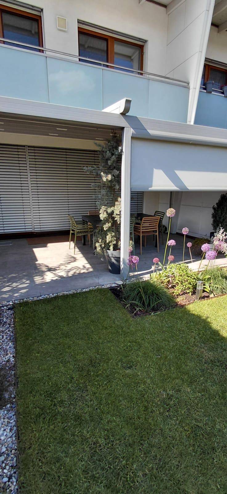 Sonniger Terrassenbereich mit Markise, Rasenfläche und blühenden Pflanzen im Vordergrund.