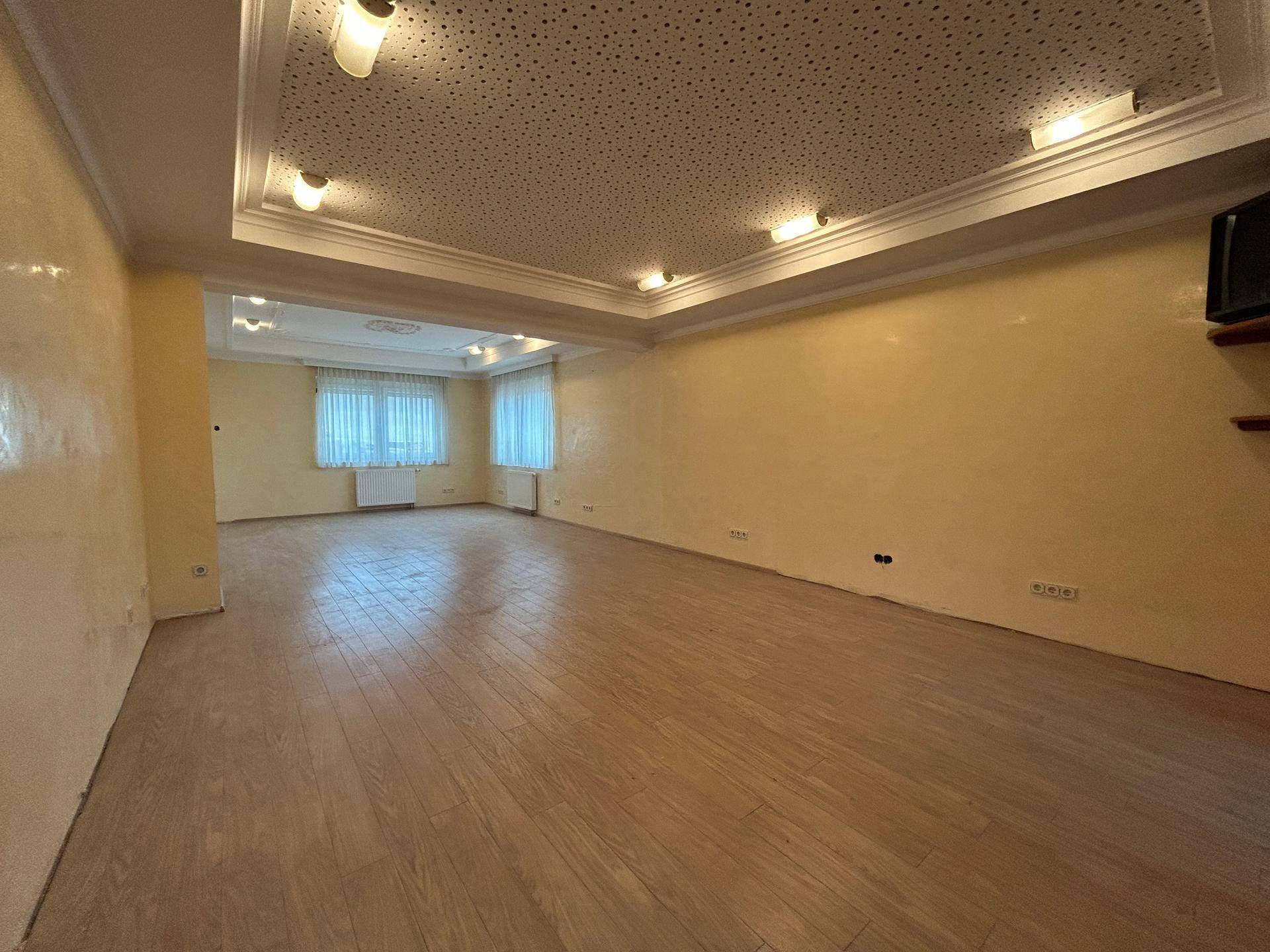 Geräumiger, leerer Wohnraum mit hellem Holzfußboden, beigen Wänden und einer dekorativen Decke mit Einbauleuchten.