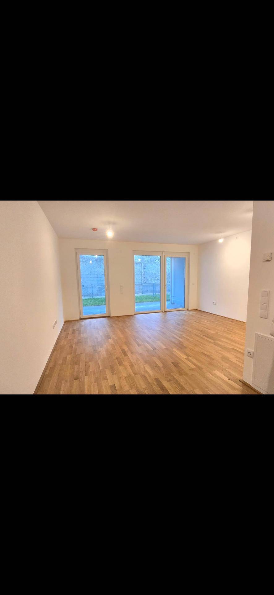 Helles, leeres Wohnzimmer mit Holzboden und Zugang zur Terrasse durch große Fenster.