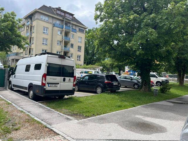Wohngebäude mit Parkplätzen und Bäumen im Vordergrund, umgeben von Grünflächen.