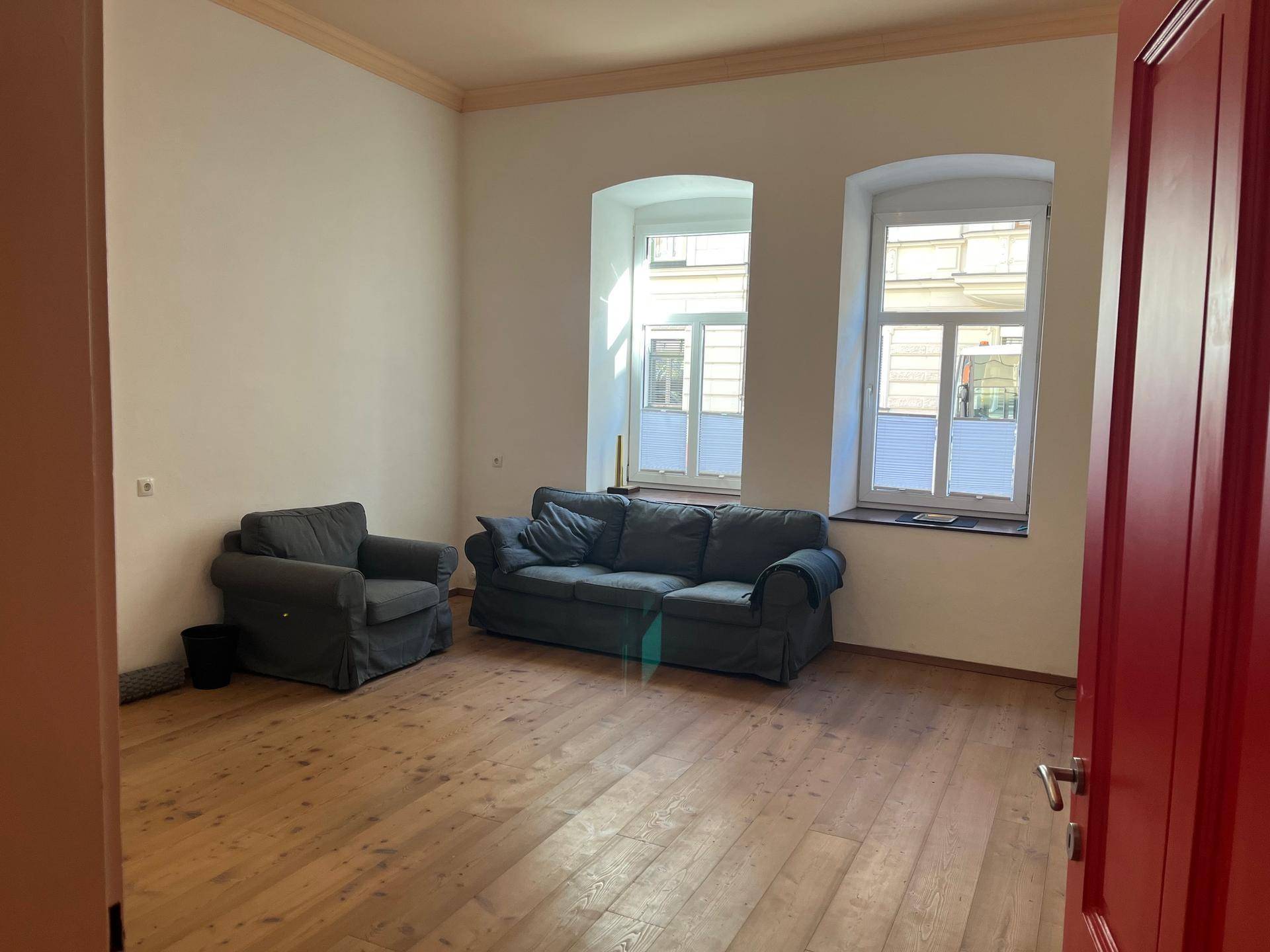 Geräumiges Wohnzimmer mit Holzdielenboden, hohen Decken und zwei großen Fenstern.