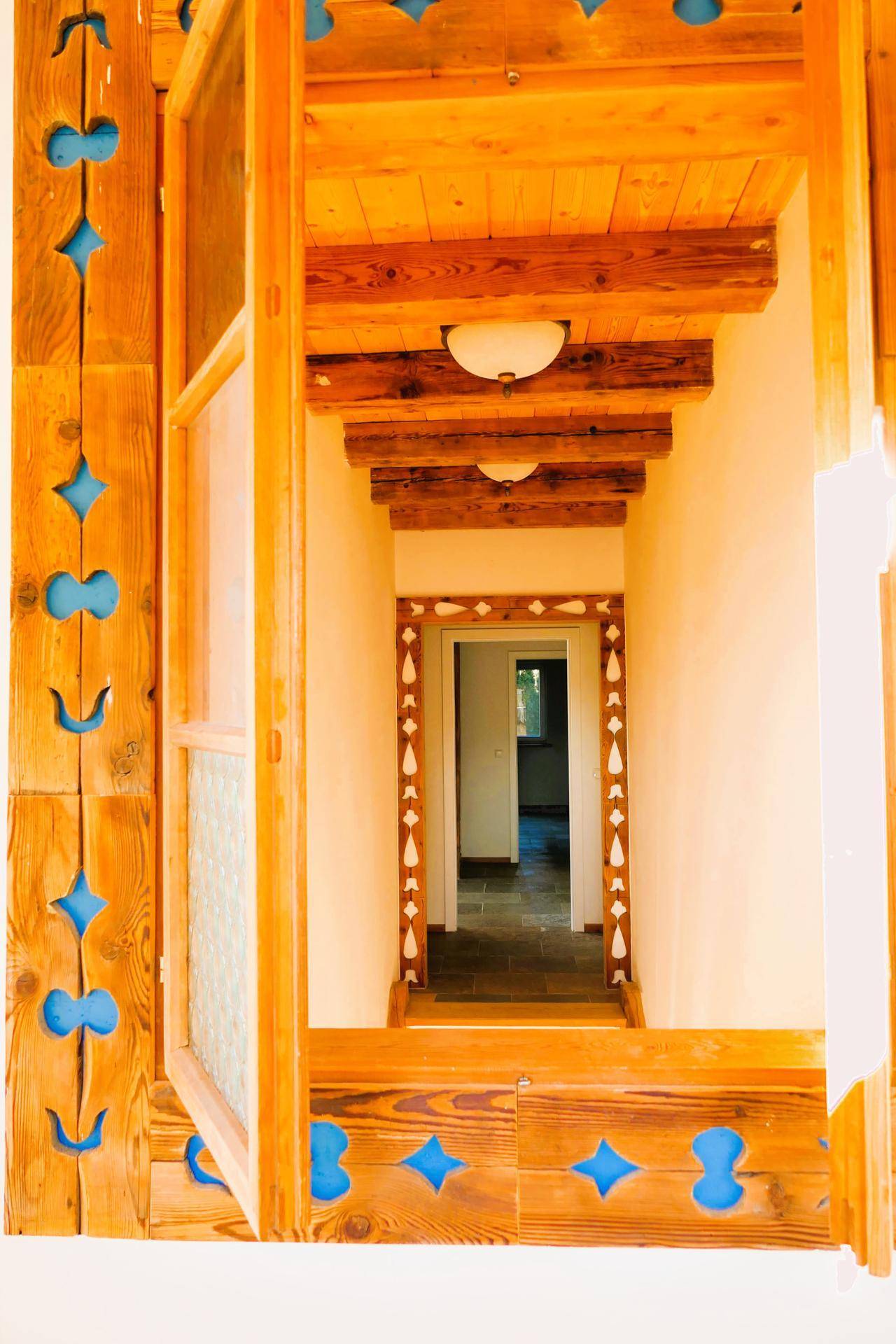 Blick durch ein kunstvoll geschnitztes Holzfenster in einen hellen Flur mit Holzbalkendecke.