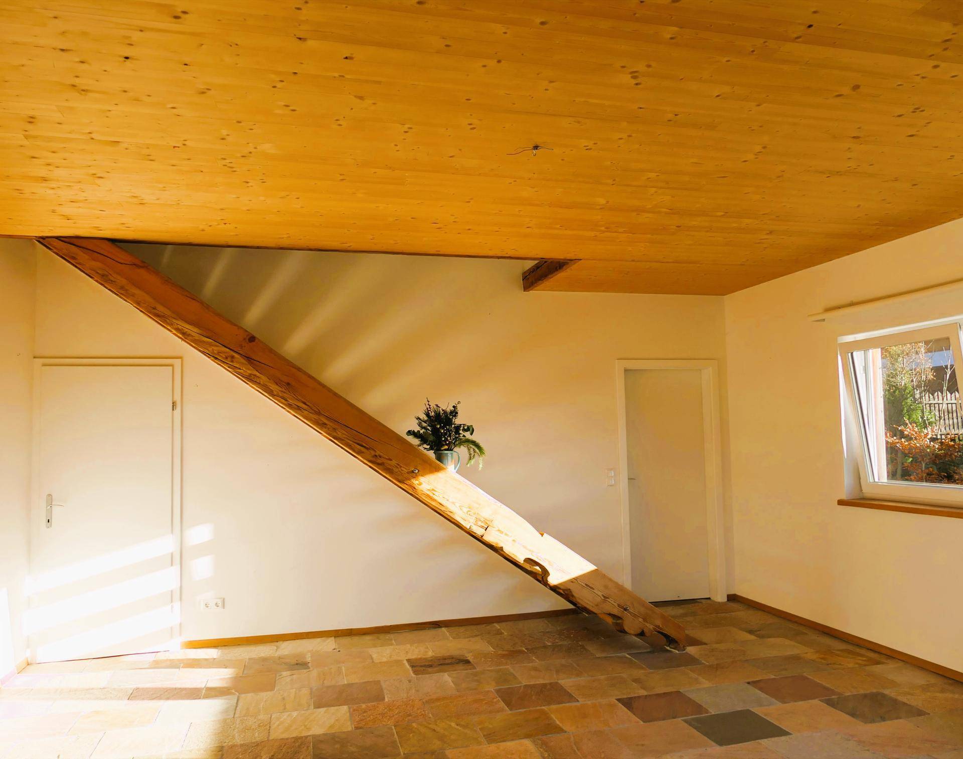 Heller Raum mit Holzbalkendecke, Fliesenboden und einer schrägen Holzkonstruktion an der Wand.