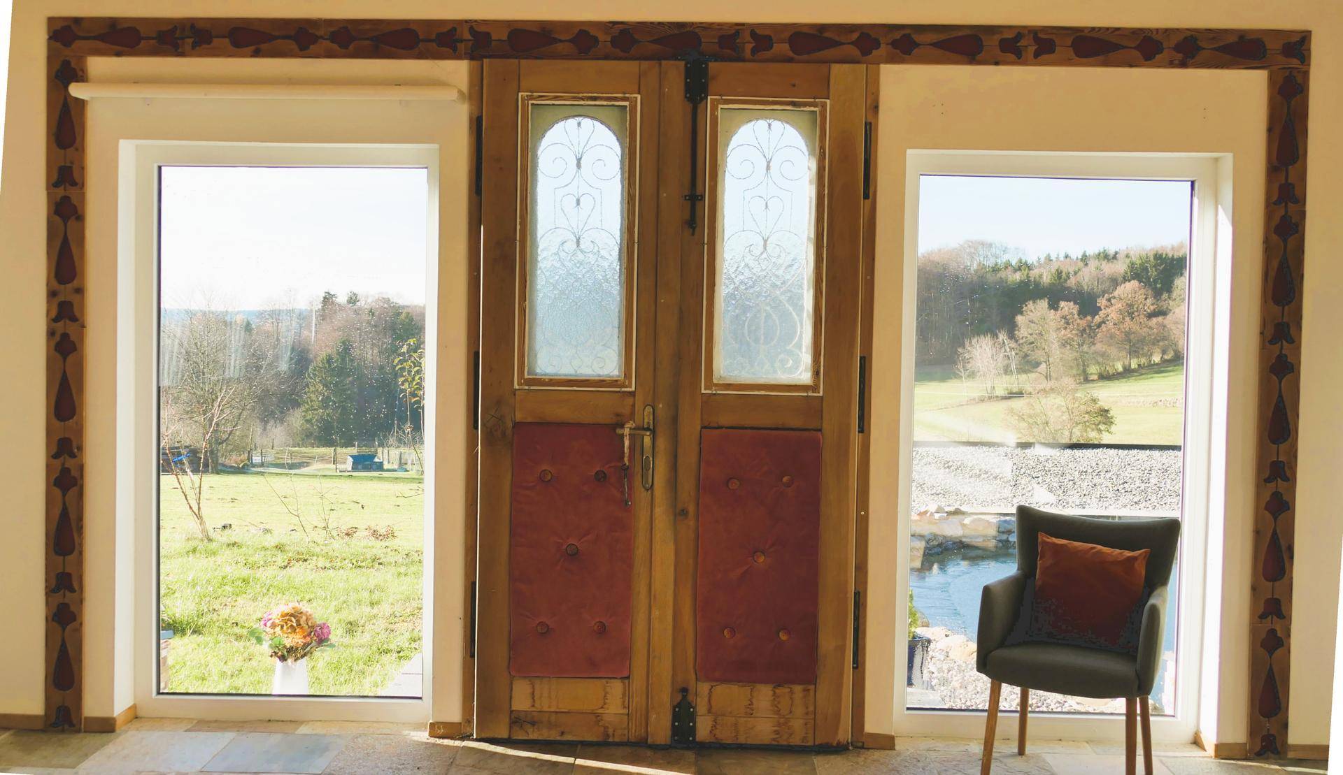Große Fenster und eine Holztür mit Blick auf die weite grüne Landschaft und den Himmel.