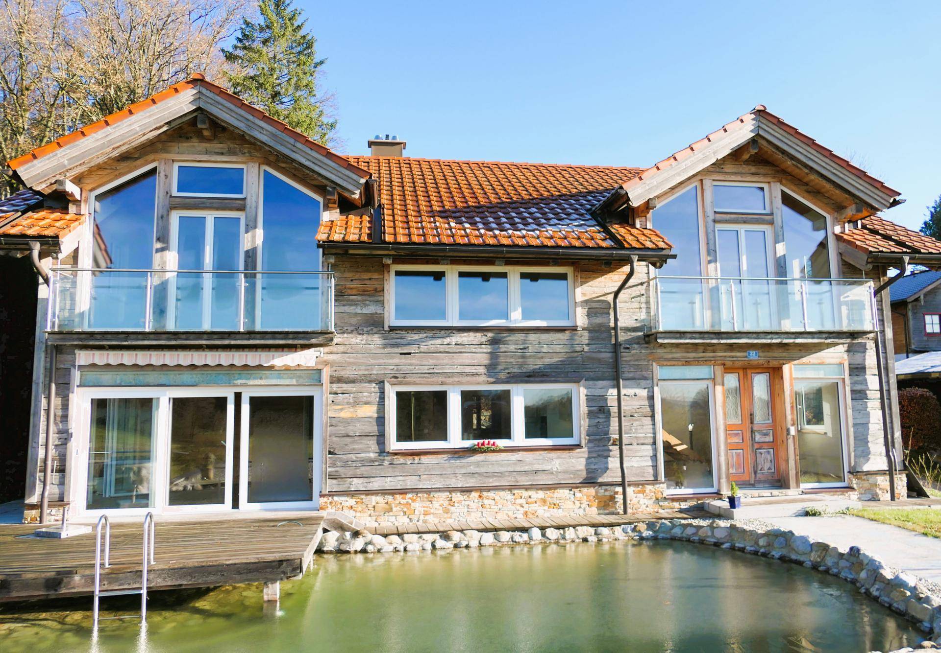 Modernes Holzhaus mit Balkonen und einem Teich im Vordergrund, umgeben von Natur.