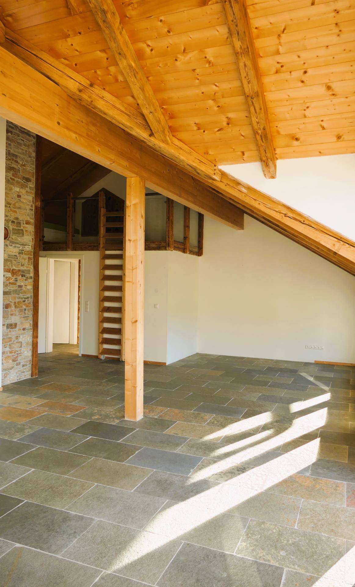Geräumiger Raum mit einer Steinwand, Holzbalkendecke und Fliesenboden, durchflutet von Tageslicht.