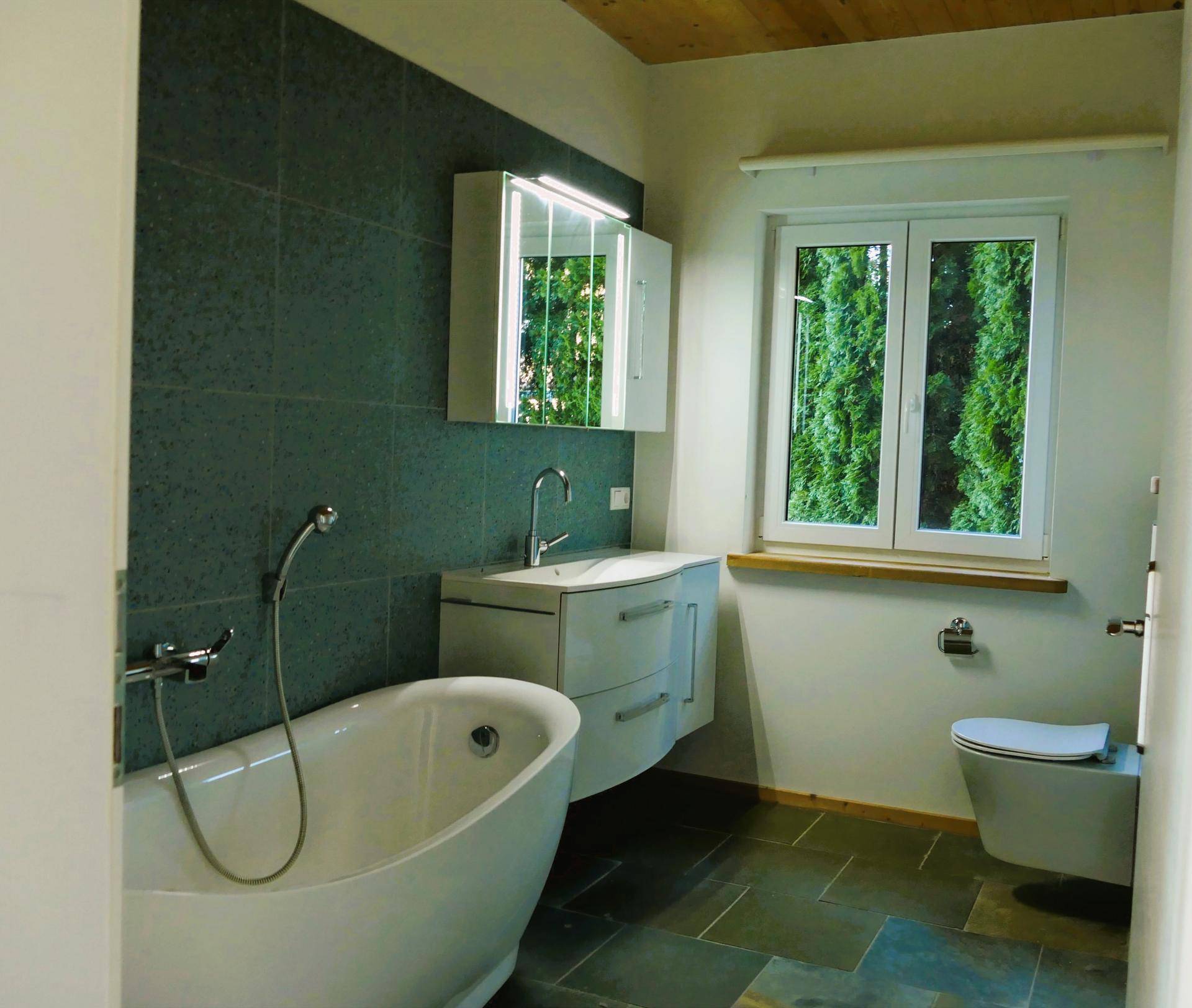 Elegantes Badezimmer mit freistehender Badewanne, modernem Waschtisch und Fenster mit Blick ins Grüne.