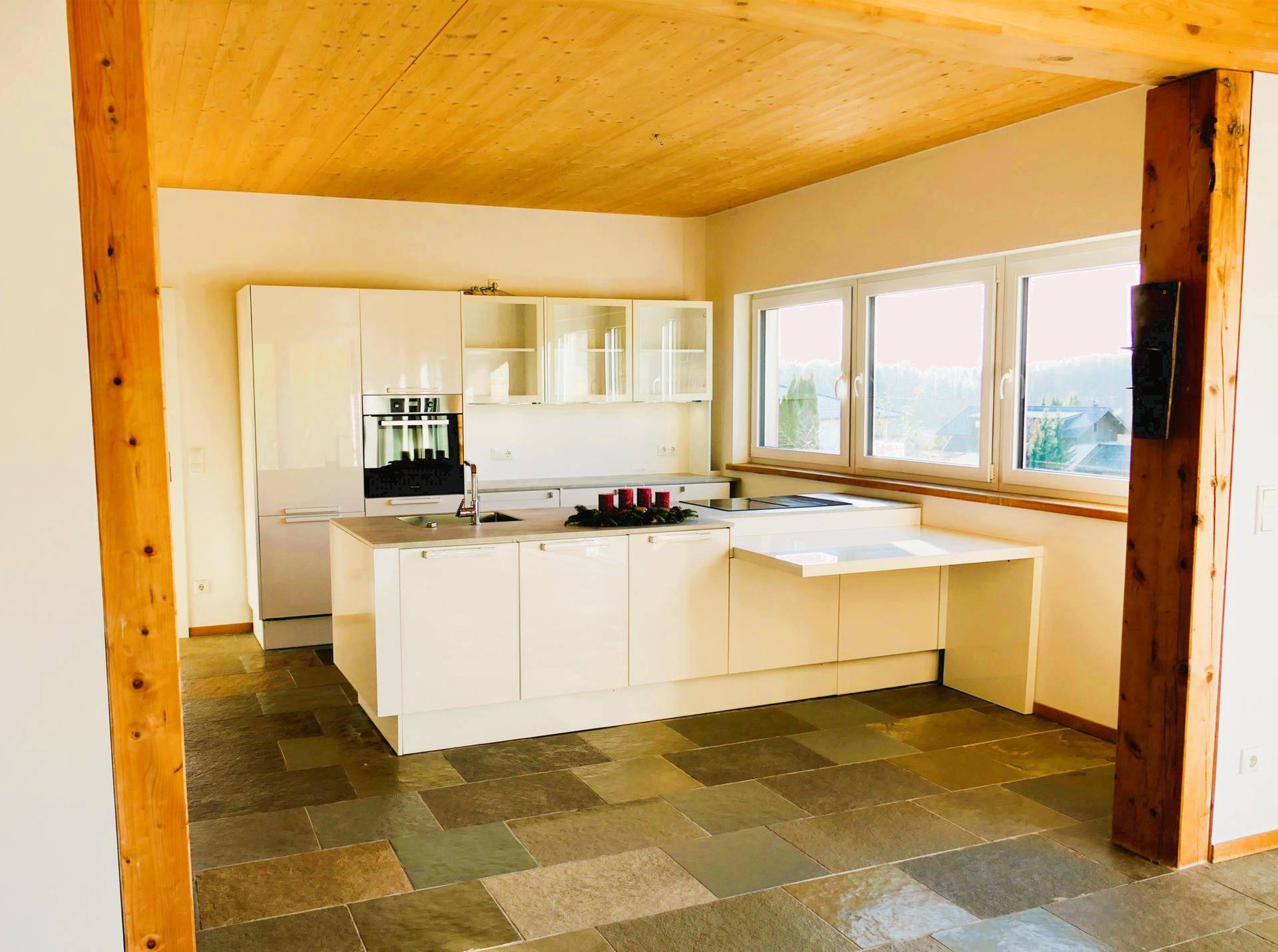 Moderne weiße Einbauküche mit Kochinsel und großen Fenstern, die viel Tageslicht hereinlassen.