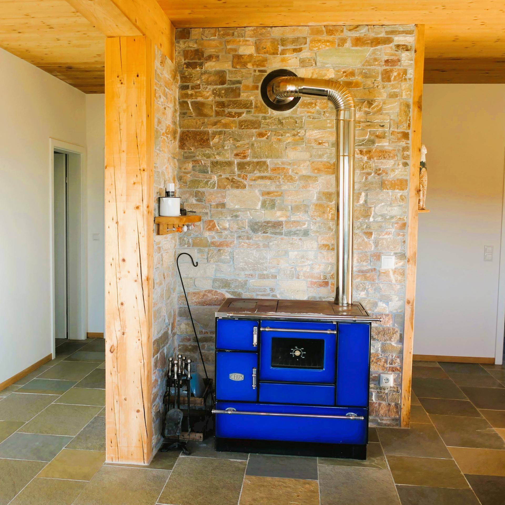 Gemütlicher Raum mit einem blauen Holzofen, Steinwand und Holzbalken an der Decke.