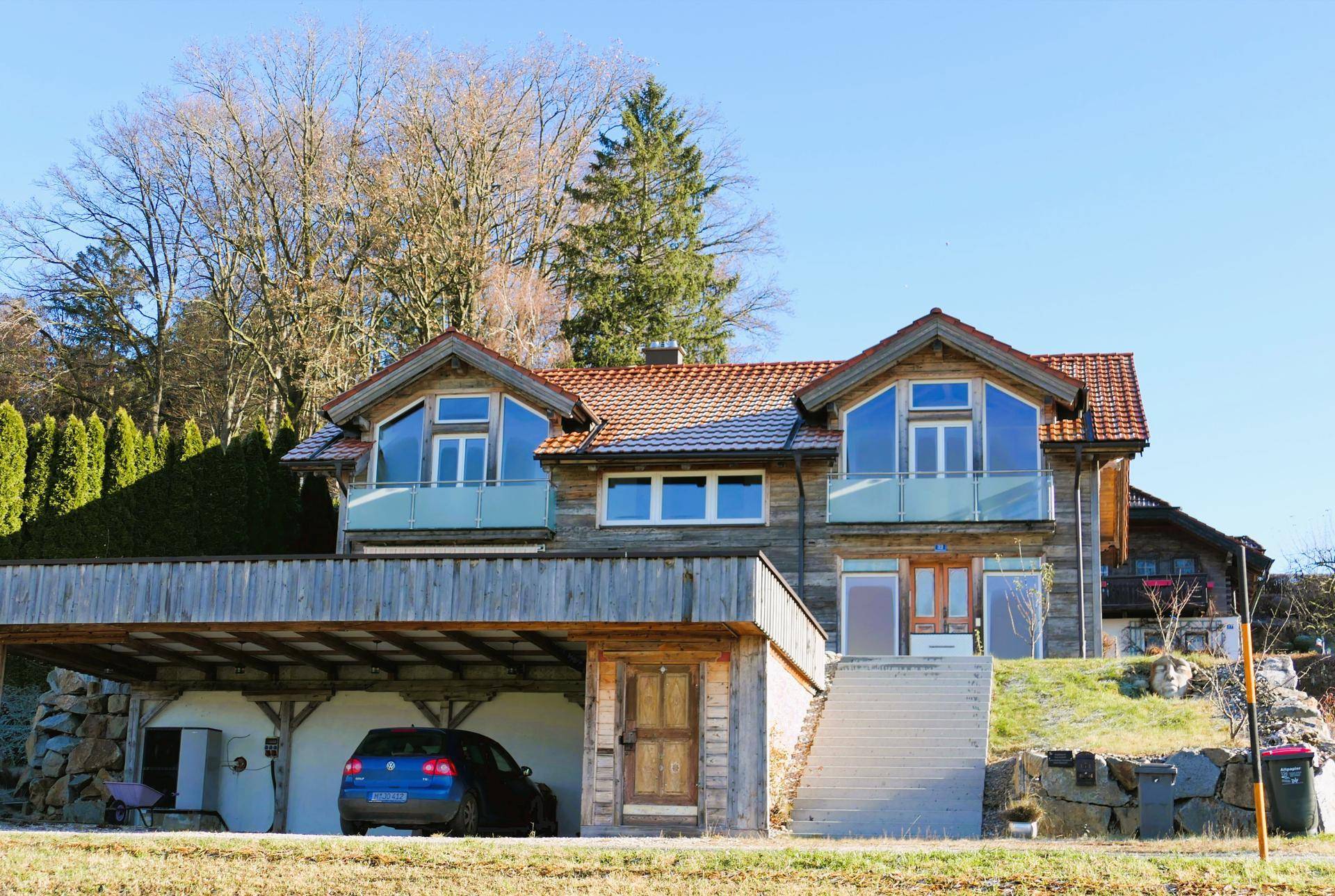 Rustikales Holzhaus mit Carport, großen Fenstern und Balkonen, eingebettet in grüne Natur.