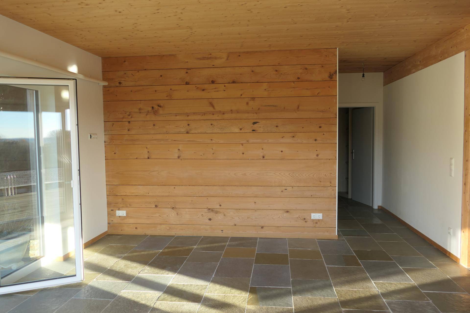 Geräumiger Raum mit einer Holzwand, Fliesenboden und großen Fenstern, die viel Licht spenden.