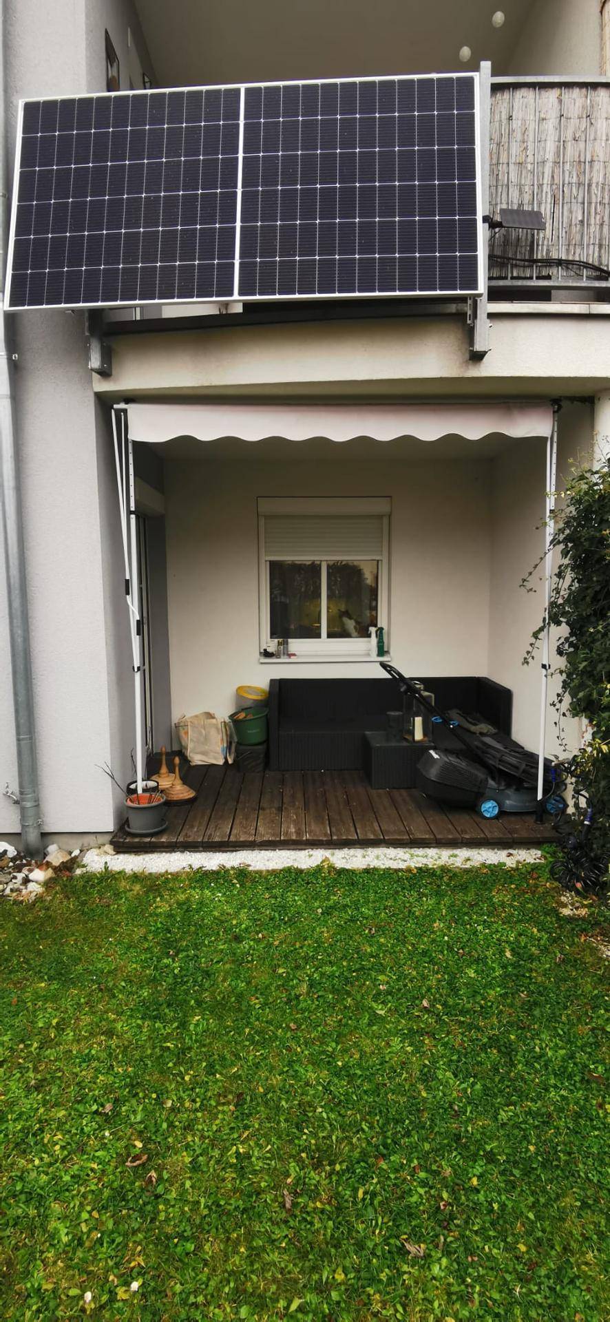 Außenbereich mit Holzterrasse, Rasenfläche und installierten Photovoltaik-Modulen über dem Sitzbereich.