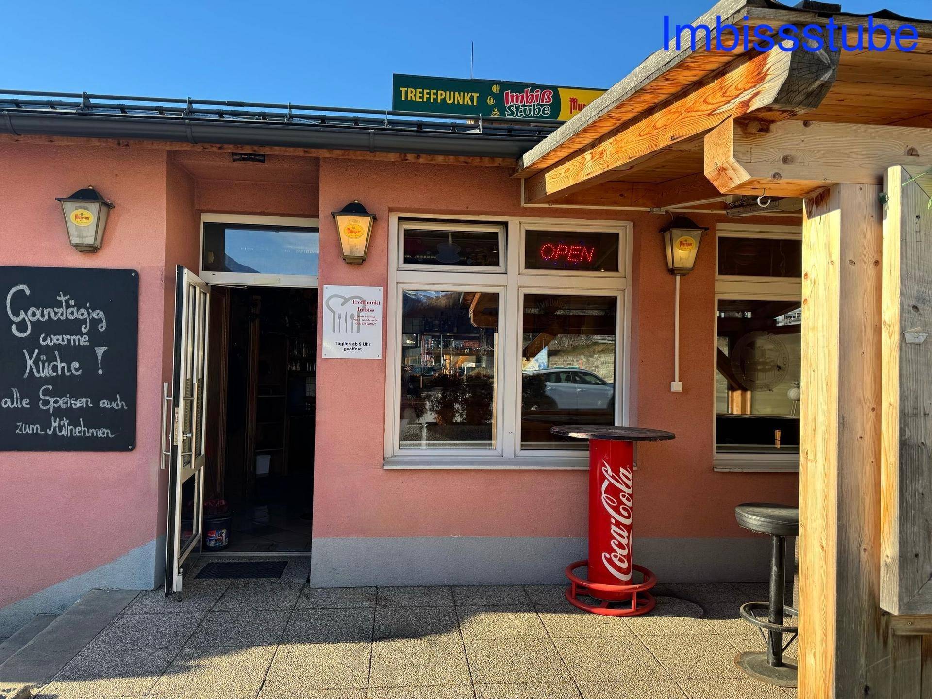 Eingangsbereich eines Imbisslokals mit offener Tür, Werbetafel und einem Coca-Cola Stehtisch.