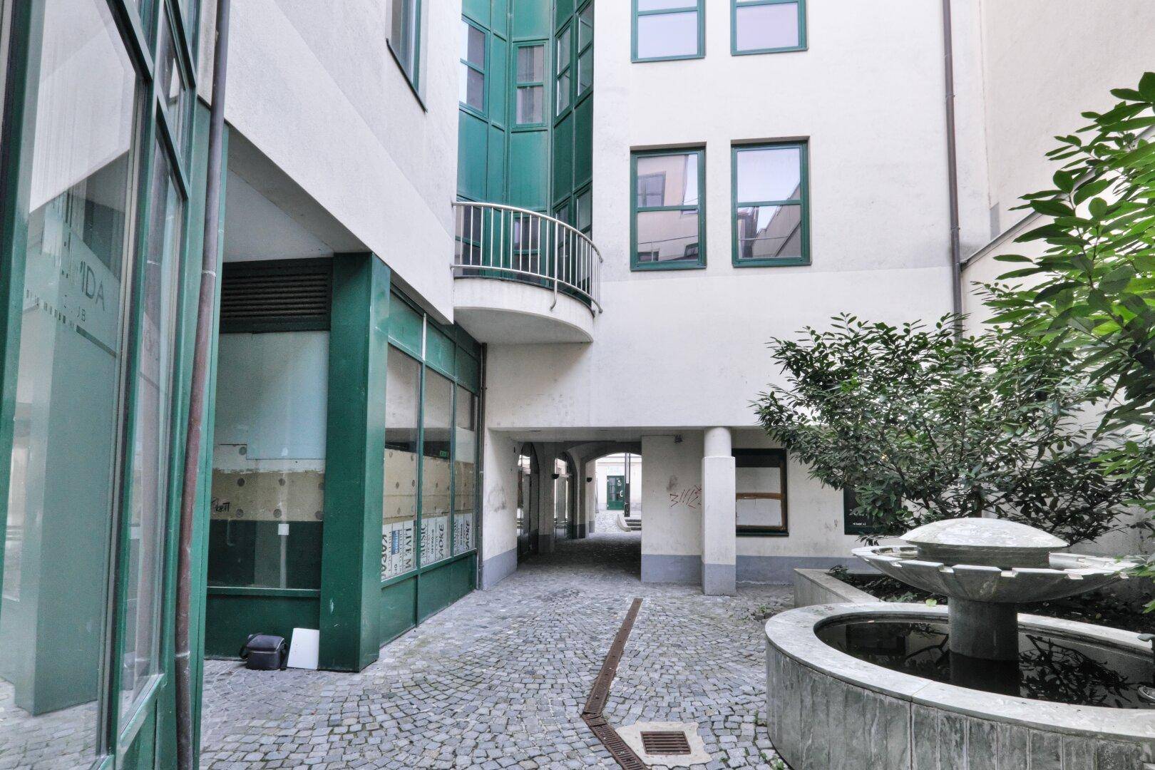 Innenhof mit Kopfsteinpflaster, einem Brunnen und begrünten Bereichen, umgeben von Gebäuden.
