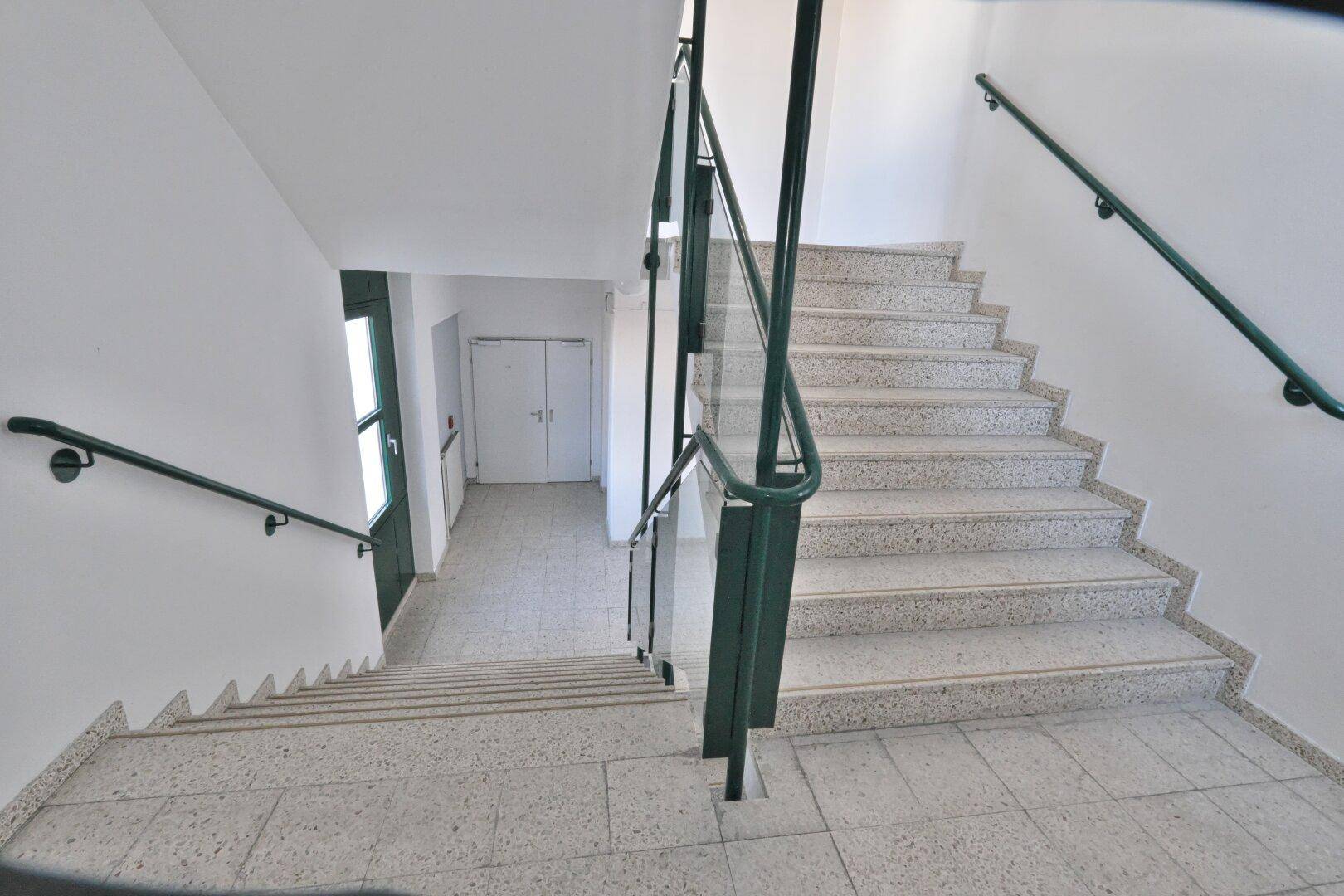 Helles Treppenhaus mit Terrazzostufen, grünen Handläufen und Fenstern für natürliches Licht.
