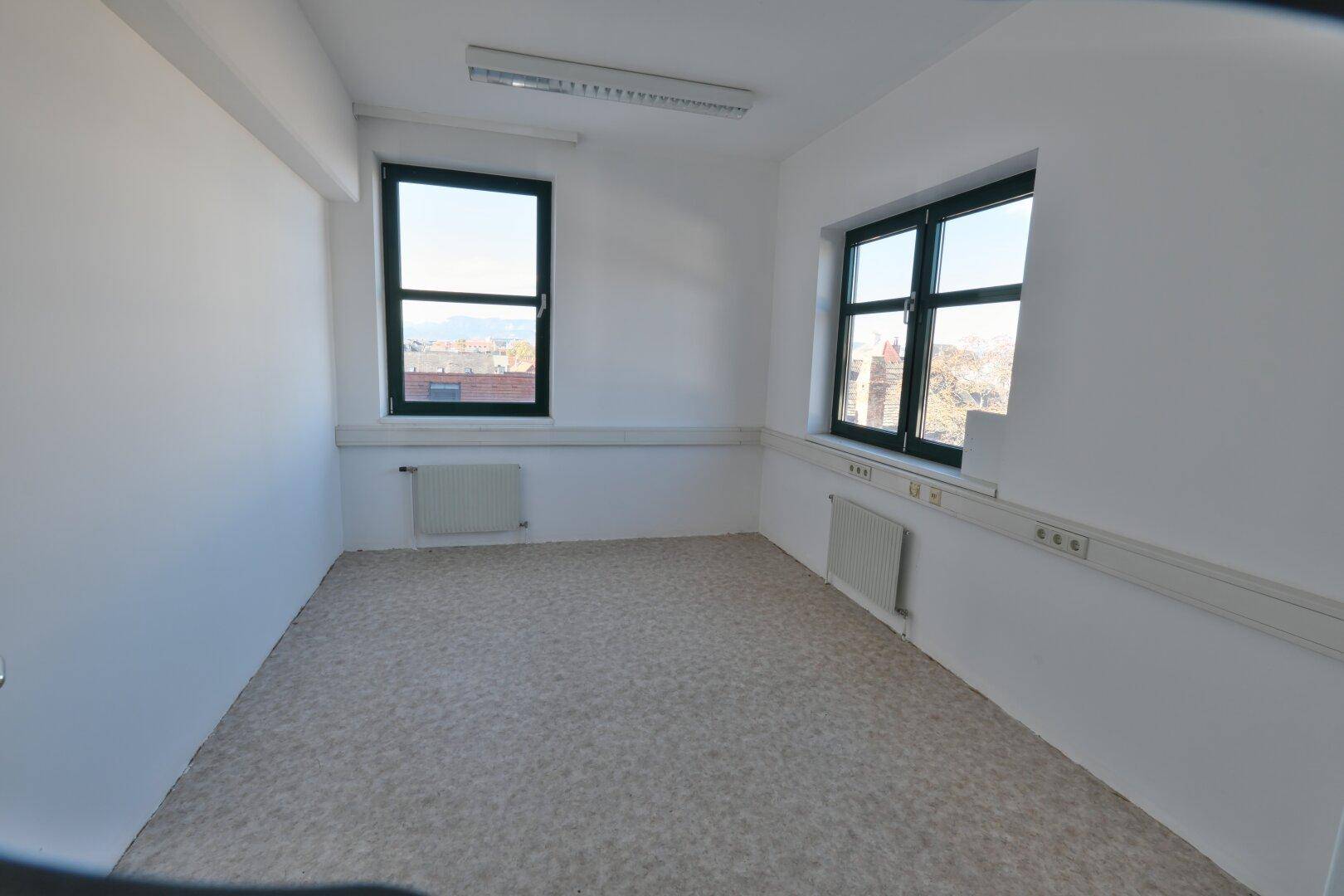 Kleinerer Büroraum mit zwei Fenstern, Teppichboden und Heizkörpern unter den Fenstern.