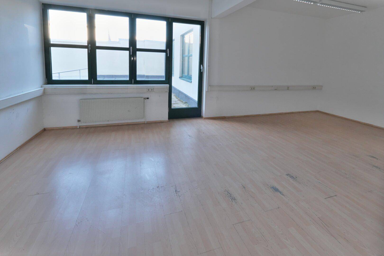 Leerer Büroraum mit hellem Laminatboden, großen Fenstern und Zugang zu einem Außenbereich.