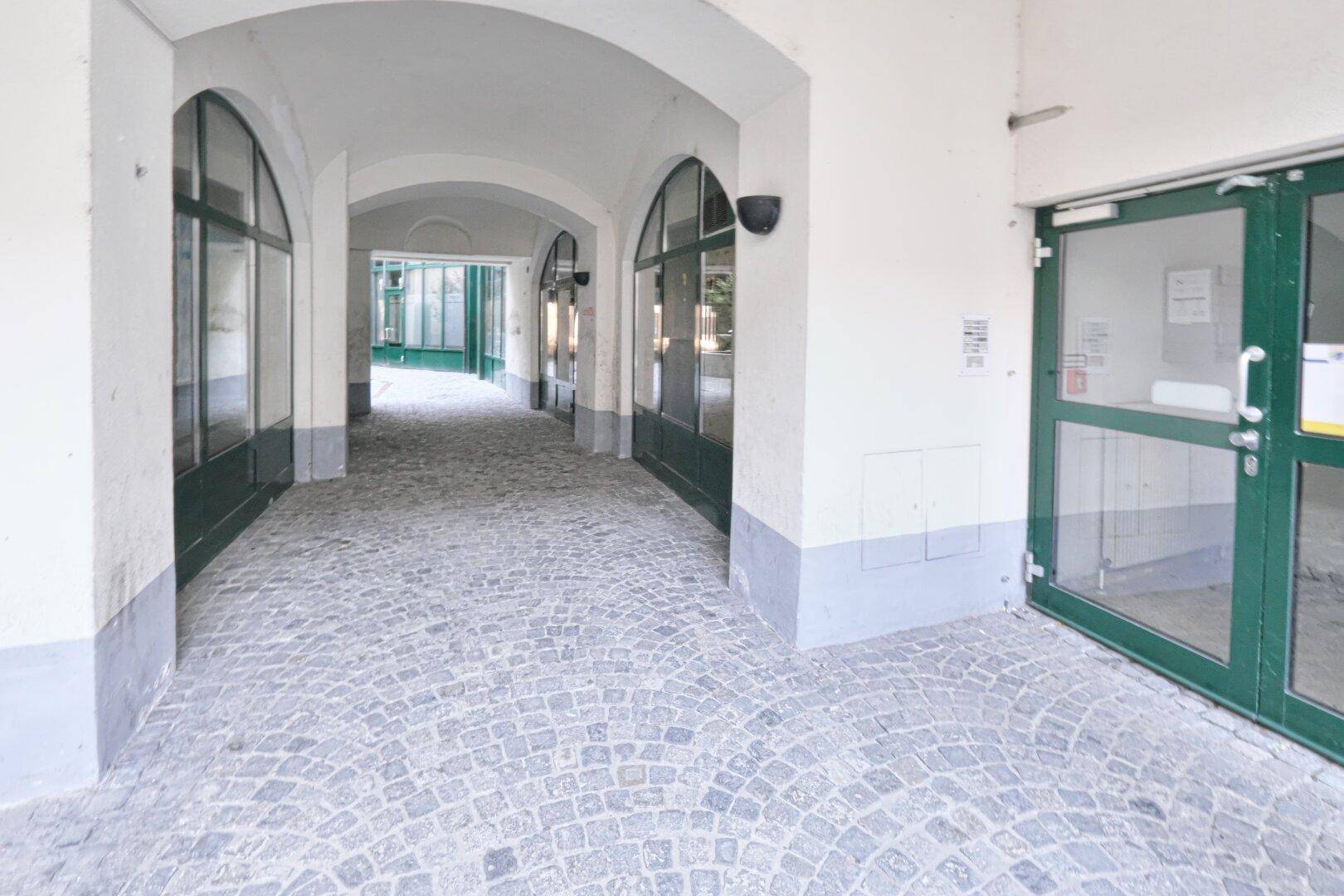 Überdachte Passage mit gewölbten Decken, Kopfsteinpflaster und Schaufenstern, die zu weiteren Bereichen führt.