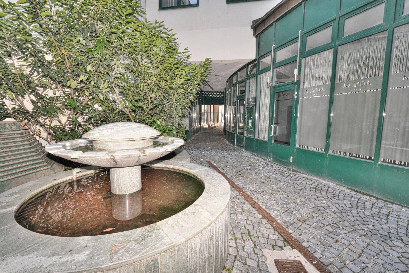 Detailansicht des Brunnens im Innenhof mit Kopfsteinpflaster und grünen Ladenfronten im Hintergrund.