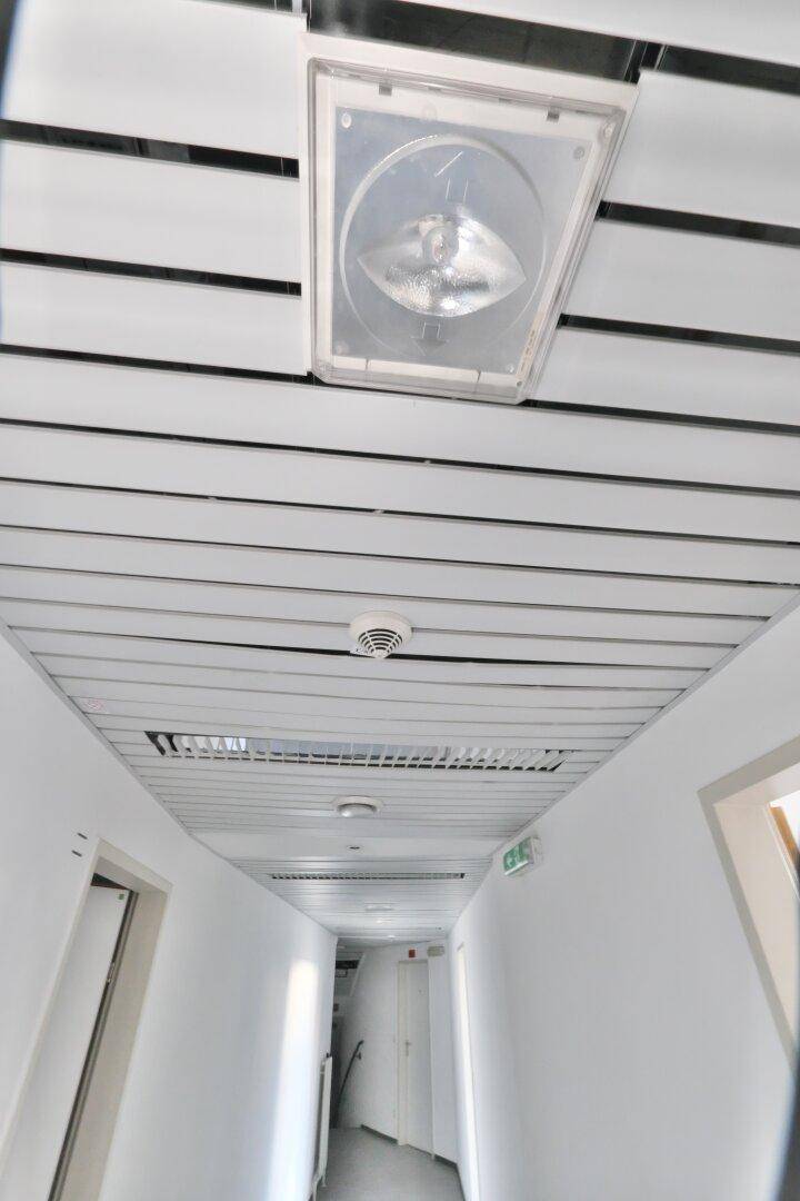 Langer, heller Flur mit abgehängter Decke, Beleuchtung und einem Rauchmelder an der Decke.