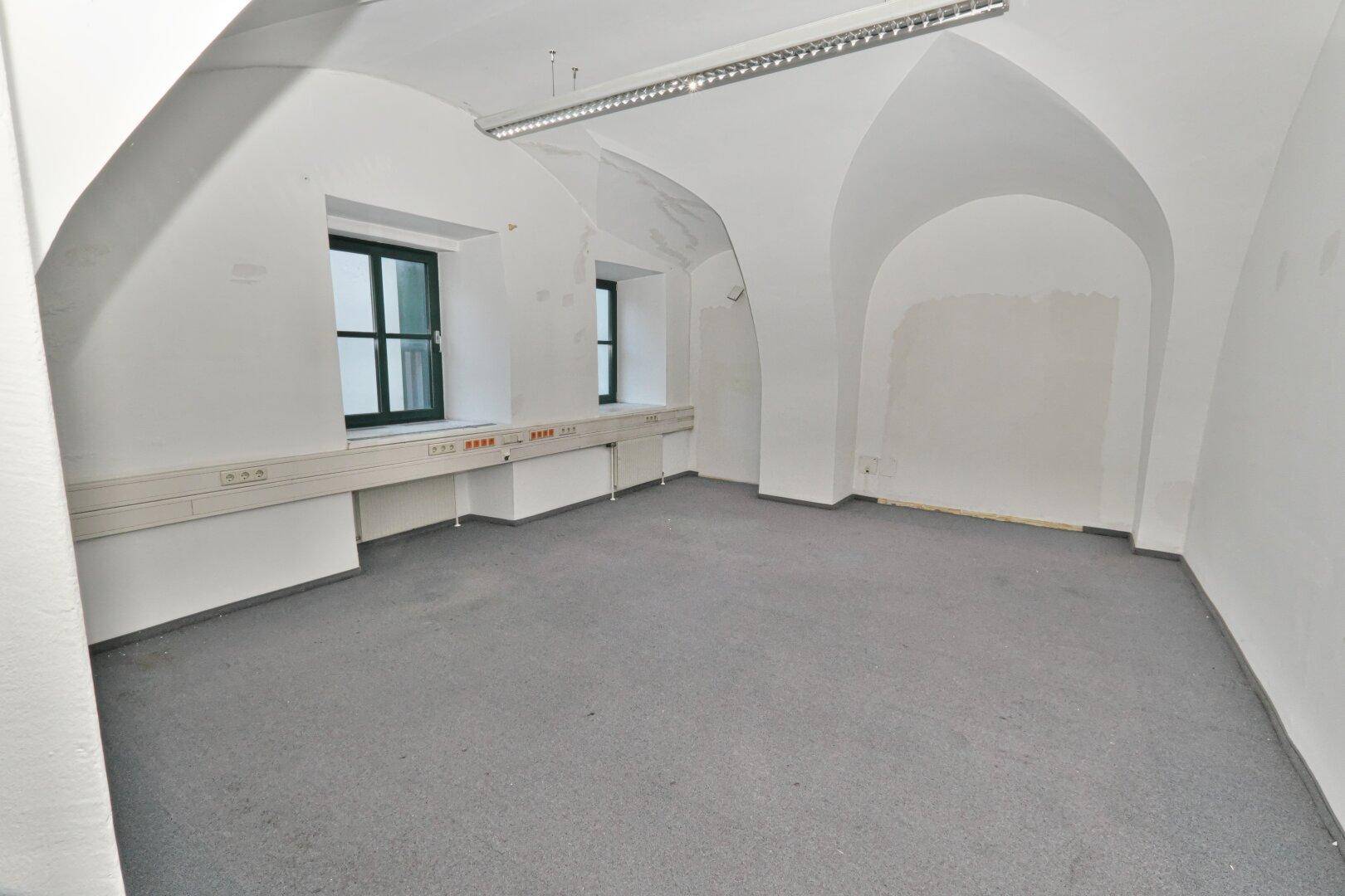 Geräumiger Büroraum mit weiß gestrichenen Wänden, gewölbter Decke und grauen Teppichboden.