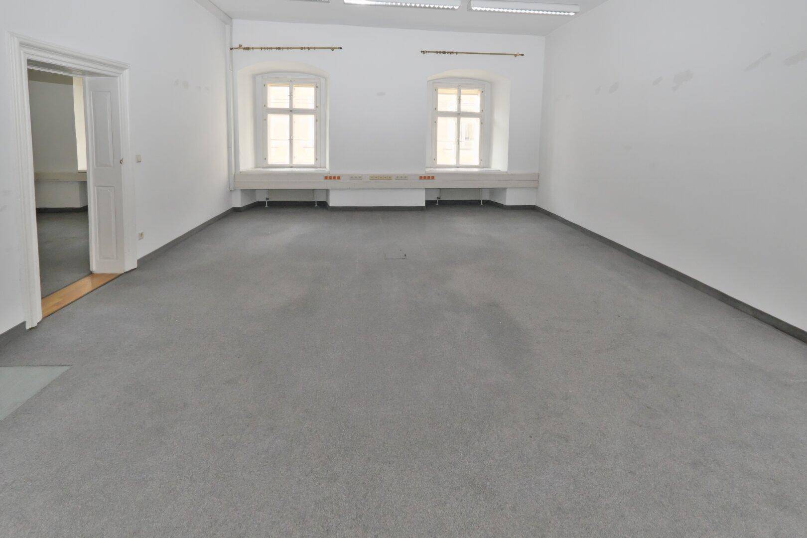 Heller Büroraum mit zwei Fenstern, weiß gestrichenen Wänden und einem grauen Teppichboden.