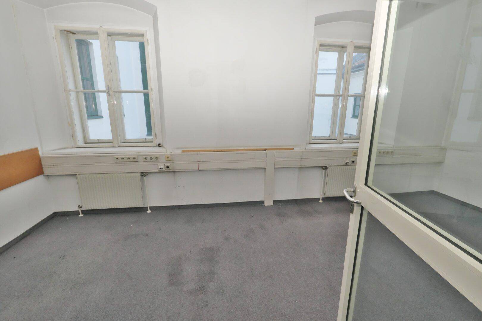 Büroraum mit zwei Fenstern, die viel Tageslicht hereinlassen, und einem grauen Teppichboden.