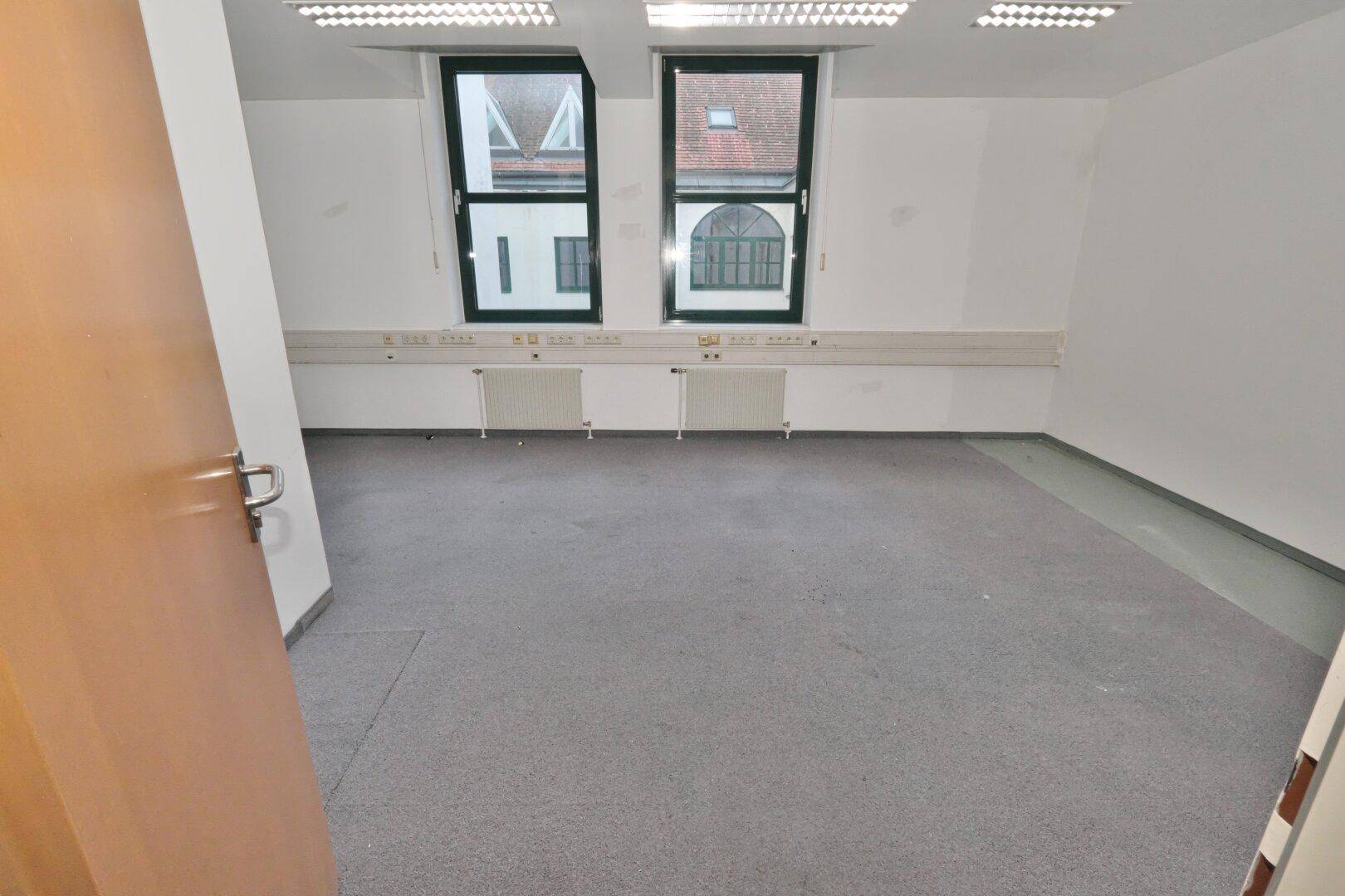 Heller Büroraum mit zwei Fenstern, grauen Teppichboden und einer langen Fensterbank mit Steckdosen.