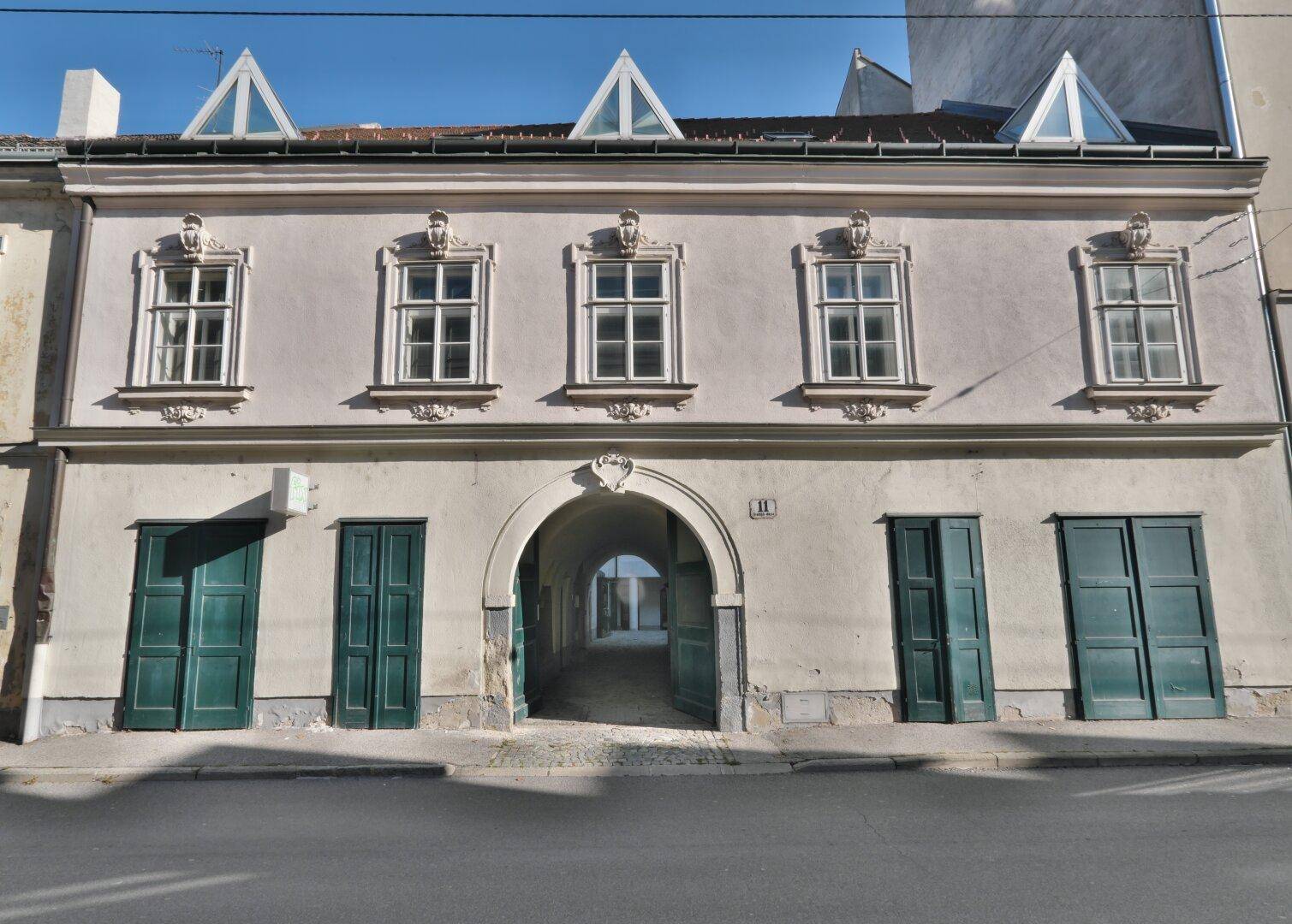 Historische Fassade eines Gebäudes mit mehreren Fenstern und einem großen Torbogen zur Straße hin.