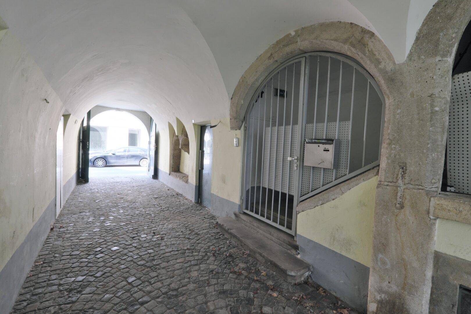 Überdachter Durchgang mit Kopfsteinpflaster und einem Gittertor, das den Zugang zu einem Treppenhaus sichert.